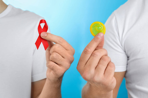 Men with condom and AIDS awareness ribbon on light blue background, closeup - Фото, изображение