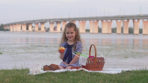 маленька усміхнена дівчинка їсть свіже яблуко і дає великий палець під час пікніка в природі біля річки
 - Кадри, відео