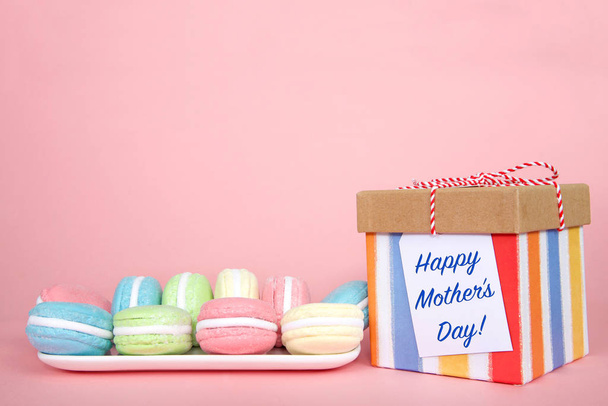 White rectangular plate with pastel colored macaron cookies laying on a pink background, colorful striped present with Happy Mother's Day note attached. - Фото, изображение