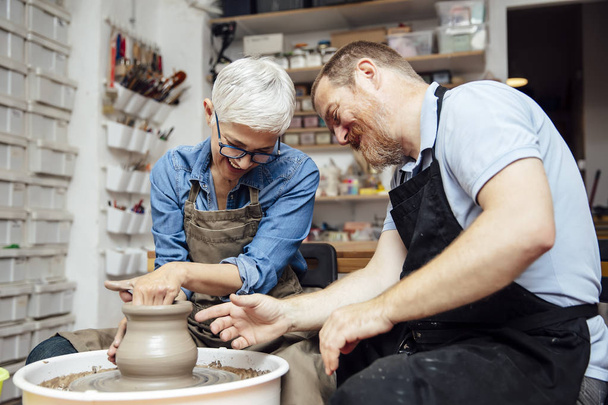 Senior woman spinning clay on a wheel with a help of a teacher at pottery class - Photo, Image