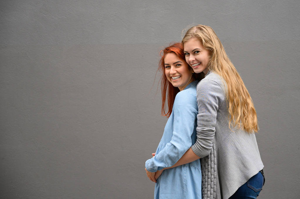 Same-sex relationships. A red-haired pregnant woman in a denim dress is standing against a gray wall, her hand is resting under her tummy. Her wife gently hugged the expectant mother from behind. - Фото, изображение
