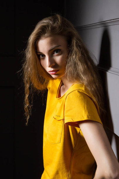 young girl in a bright yellow T-shirt stands in a dark room against the wall - Foto, imagen