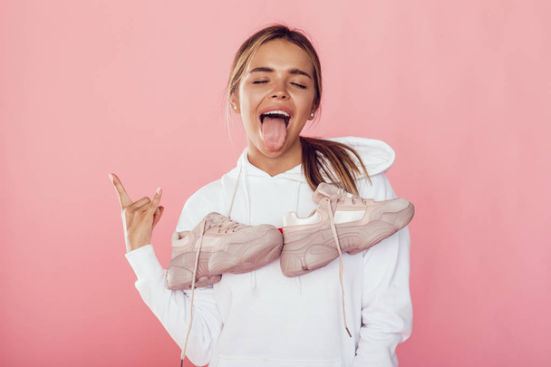 Young emotional woman shows tongue and stands with sneakers on her shoulders. - Valokuva, kuva