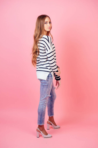 young and attractive woman stylish model posing in jeans , high-heeled shoes and a sweater in studio on a pink background - Photo, Image