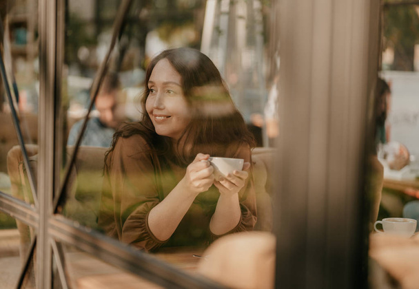 Affascinante donna bruna con lunghi capelli ricci seduta alla finestra in caffè con tazza di caffè in mano
 - Foto, immagini