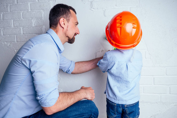 happy family handsome father and charming cute little son in helmet hardhat painted wall with paint white color . concept of repairs house apartment by young family - Foto, Bild