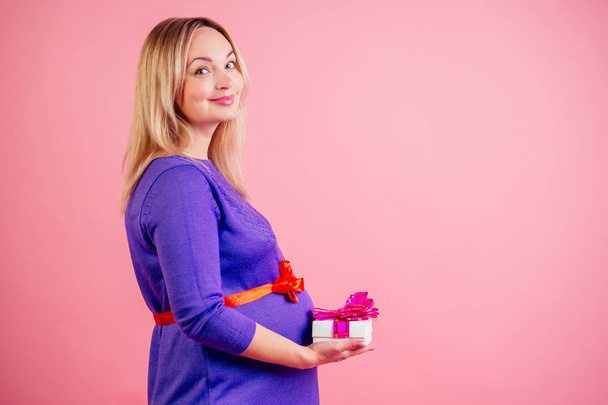 charming and gentle blonde pregnant woman belly baby bump hold a box with a gift in a dress in studio on a pink background . concept of baby shower - Foto, imagen