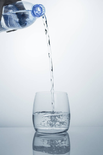 Ένα υψηλό ρεύμα καθαρού πόσιμου νερού ρέει από το μπουκάλι σε ένα διαυγές ποτήρι. - Φωτογραφία, εικόνα