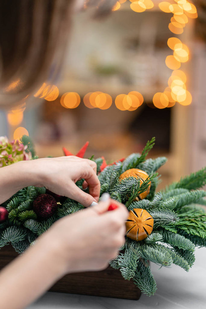 Νεαρή γυναίκα δημιουργεί και διακοσμεί όμορφη εορταστική ρύθμιση από φρέσκο έλατο, καλλωπιστικά φυτά σε ένα ρουστίκ ξύλινο κουτί. Χριστουγεννιάτικη διάθεση. Γιρλάντα bokeh στο παρασκήνιο. - Φωτογραφία, εικόνα