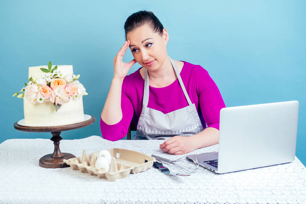 confectioner housewife pastry-cook businesswoman looks disappointed at the white two-tiered wedding cake with fresh flowers on a table in studio on a blue background - Photo, image