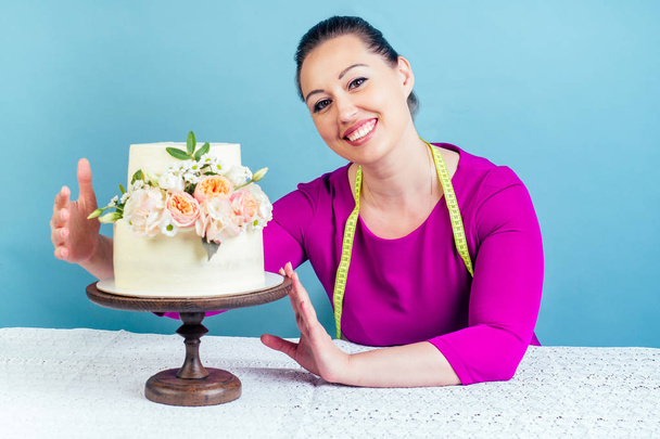 hungry and funny housewife confectioner fat woman hugs appetite a high-calorie wedding cake with measuring tape around the neck in studio on a blue background. concept of diet - 写真・画像