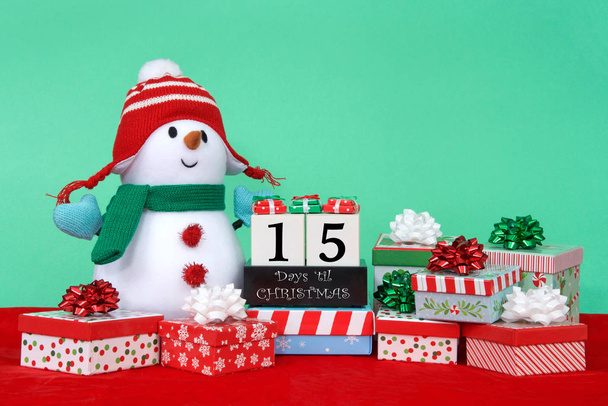 Töltött hóember zöld sállal, kék kesztyű, piros és fehér kalap ül a piros bársony körül fényes színes ajándékok, blokkok visszaszámolni karácsonyig. 15 nap - Fotó, kép