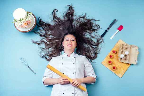 top view from above of confectioner pastry chef baker woman with long dark hair around wedding birthday cake , cookies eggs, whisk and rolling pin lying on the floor in studio on blue background. - Valokuva, kuva