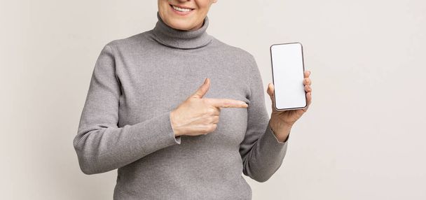 Mujer mayor mostrando la pantalla del teléfono inteligente en blanco y señalando en él
 - Foto, Imagen