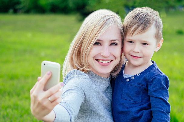 attractive mother and loved son take pictures selfie on phone in the park on a background of green grass and trees - Φωτογραφία, εικόνα