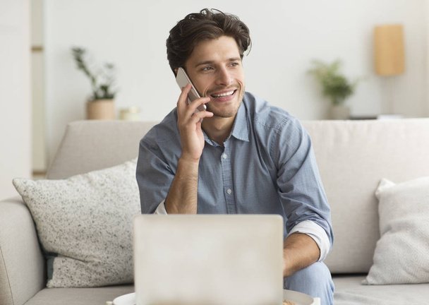 Młody człowiek o przyjemnej rozmowy telefonicznej siedzi na laptopie w domu - Zdjęcie, obraz