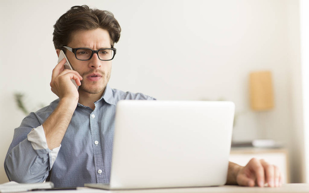 Poważny facet mówiący przez telefon komórkowy pracujący na laptopie w biurze - Zdjęcie, obraz