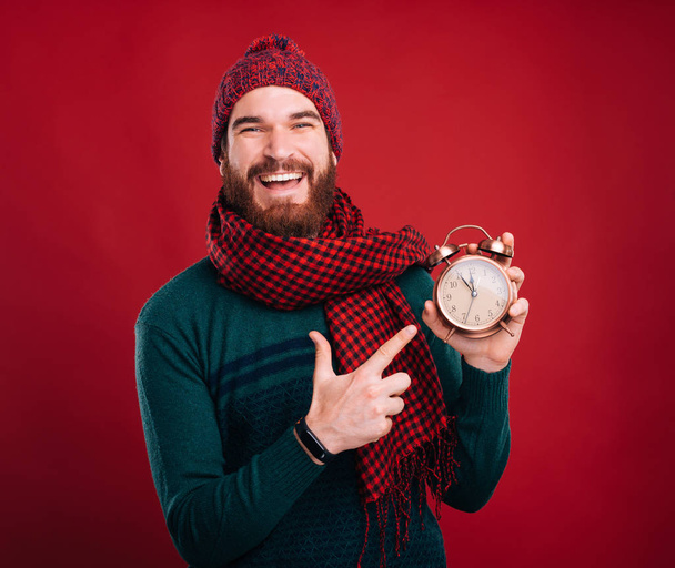 fröhlicher junger Mann in Winterkleidung zeigt auf Wecker - Foto, Bild