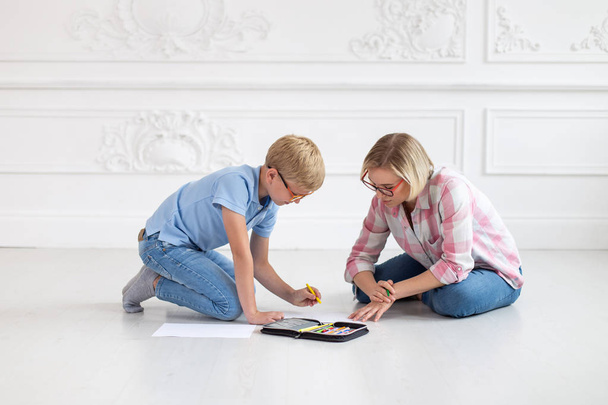 A young tutor in a plaid shirt and her young student, wearing a blue t-shirt and glasses, sit on a white floor. The woman watches as the boy draws - Foto, immagini
