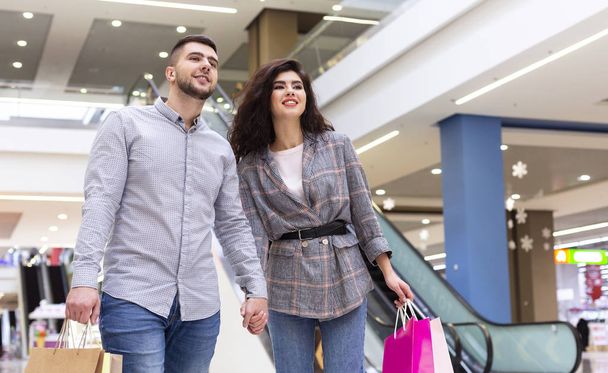 Χαρούμενο ζευγάρι με χάρτινες σακούλες που περπατούν στο εμπορικό κέντρο - Φωτογραφία, εικόνα