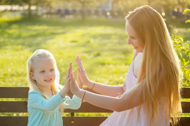 cute little blonde girl daughter in a blue dress sitting on a bench with beautiful long-haired mother playing and having fun in the summer park. happy motherhood - Φωτογραφία, εικόνα