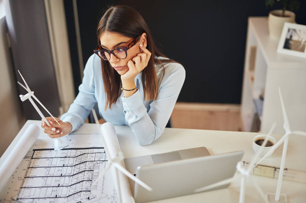 Urocza pracowity brunetka ubrana w formalne zużycie siedzi na biurku w biurze, trzymając model wiatraka i patrząc na plany. Na biurku jest laptop. - Zdjęcie, obraz