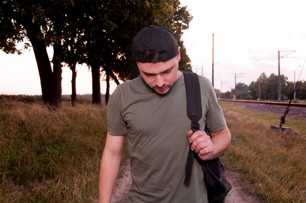 Ένας νεαρός με το κεφάλι σκυμμένο στο δρόμο με θλίψη στο πρόσωπό του. Ο τύπος με την κατάθλιψη περπατάει κατά μήκος του δρόμου κατά μήκος των δέντρων.. - Φωτογραφία, εικόνα