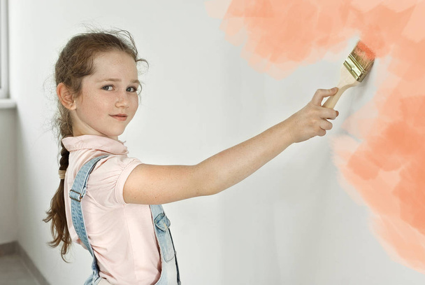 Little girl makes repairs in blue overalls. The child paints the walls with pink paint. Girl with a brush in her hands on a painted wall background. - Foto, Imagem