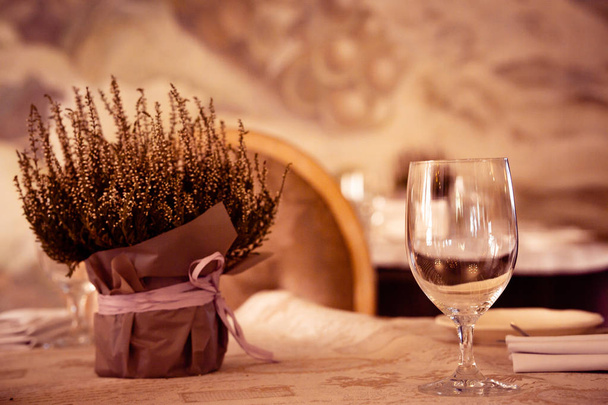 Heather bloemen in een vaas gewikkeld met licht papier. Er is een leeg wijnglas in de buurt. Op een tafel bedekt met een tafelkleed en gekleed met servetten. Warme toon. Horizontale oriëntatie. - Foto, afbeelding