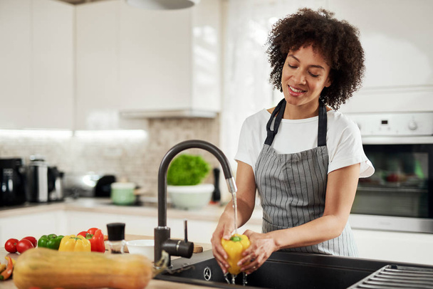 Charming mixed race woman in apron standing in kitchen ad washing yellow paprika in sink. On kitchen counter are different sorts of vegetables. - Photo, Image
