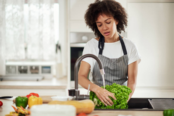 Beautiful mixed race woman with curly hair and in apron standing in kitchen and washing salad in sink. - Foto, afbeelding