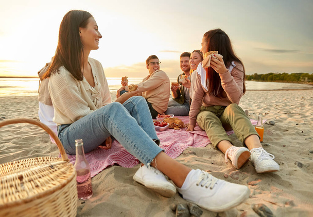 happy friends eating sandwiches at picnic on beach - Fotó, kép