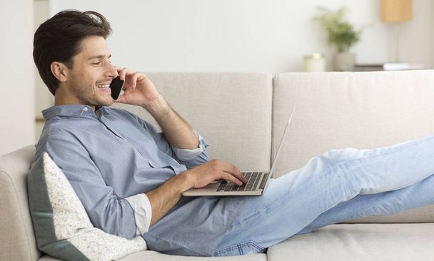 Θετικός άνθρωπος έχοντας τηλεφωνική συνομιλία που βρίσκεται με το laptop στον καναπέ - Φωτογραφία, εικόνα