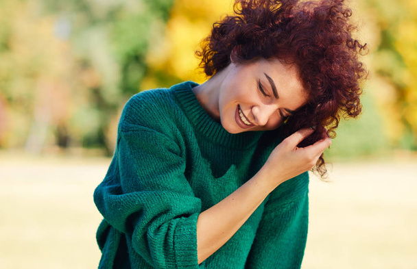 Happy beautiful young woman smiling with hand on the hair, against nature background with windy curly hair, have positive expression, dressed in green sweater. People, travel and lifestyle concept.  - 写真・画像