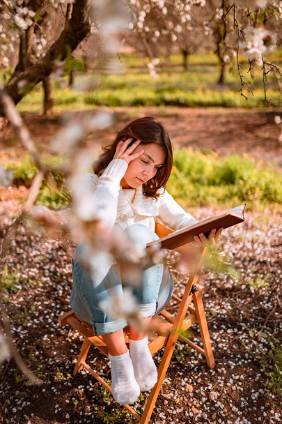 Nuori kaunis nainen valkoisessa villapaita istuu puinen tuoli alla kukkivat kolme ja lukea kirjaa
 - Valokuva, kuva