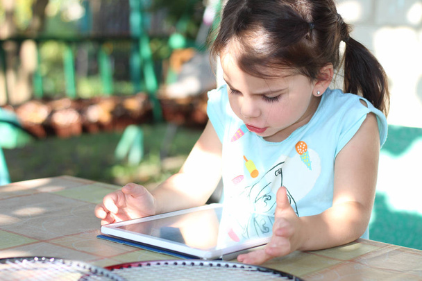 chica blanca con coletas se sienta en una mesa al aire libre y mira a la tableta
 - Foto, Imagen