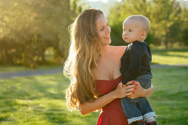 caring and beautiful long-haired woman in cocktail red dress holding the baby boy son in her arms in the park in summer .love and care concept - Foto, Bild