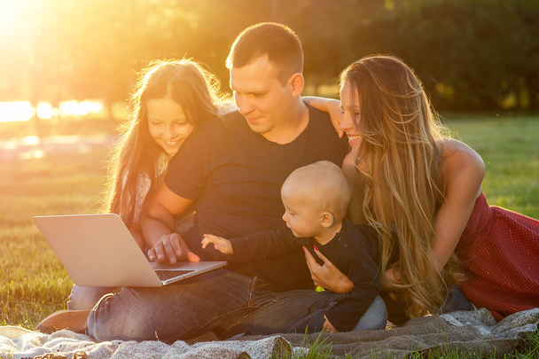 little baby boy together with his father and beautiful daughter next to charming mother play and develop looking into laptop on a blanket in the park in summer. happy family resting outdoors - Photo, image