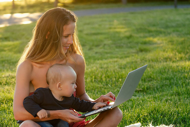 szülési szabadság ötlet. bájos és vonzó gyönyörű nő egy baba fia dolgozik a szabadban a nyári parkban laptop ül a füvön gyep zöldek - Fotó, kép