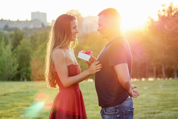 lover boyfriend gives a gift box to embarrassed attractive beautiful blonde long hair bride in love in evening cocktail dress in summer park background. An offer of marriage romantic concept - Photo, image