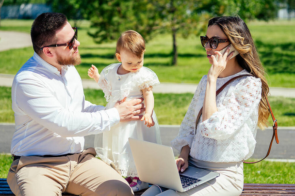 beautiful young businesswoman with chic long curly hair working on a laptop sitting on a bench next to a husband and a little daughter in white dress . rich family is resting in the park in the summer - Фото, изображение