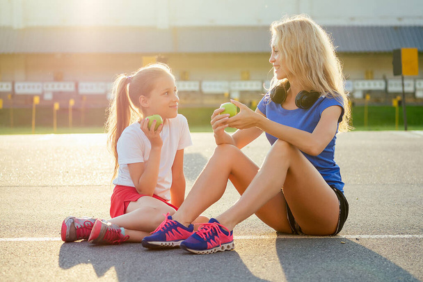 young and beautiful blonde woman together with a little girl in sportswear sitting on the asphalt and eat a green apple at the stadium. Active family mother and daughter jogging in the summer morning - Photo, Image