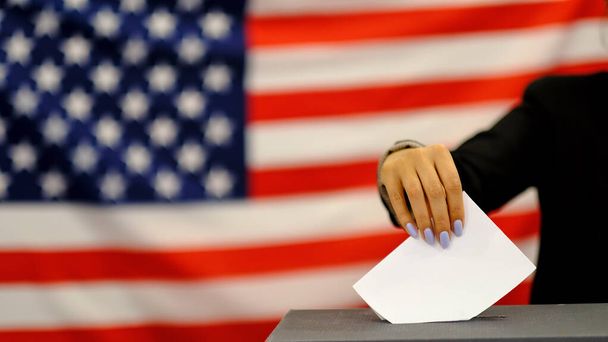 選挙日に投票箱に投票用紙を入れる女性。USAフラグの背景に白い票の紙で手のクローズアップ. - 写真・画像