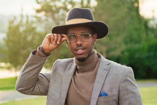 a young and handsome stylish model afro- american man in a stylish brown suit and a black hat put on glasses in a summer park . latino american hispanic businessman black guy photo model - Zdjęcie, obraz