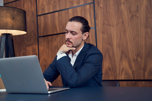 Photo on side of young businessman sitting at table with laptop on background of wooden wall . - Foto, immagini