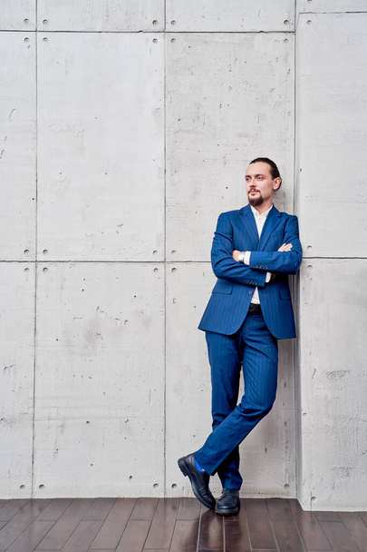 Full-length photo of young brunet in business suit with arms crossed standing near concrete wall - Foto, afbeelding