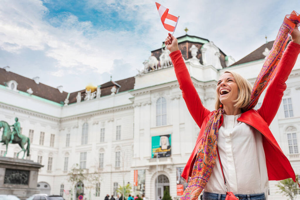 Woman tourist stands against the backdrop of the Austrian National Library in Vienna with raised arms and the flag of Austria, Vienna - Foto, Imagem