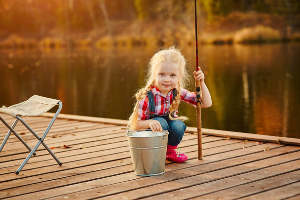 Little girl fisherman with a fishing rod and a bucket of fish on a wooden pier. - Фото, зображення