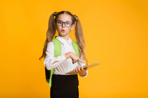 Confused little schoolgirl doing homework on digital tablet, yellow background - Photo, Image
