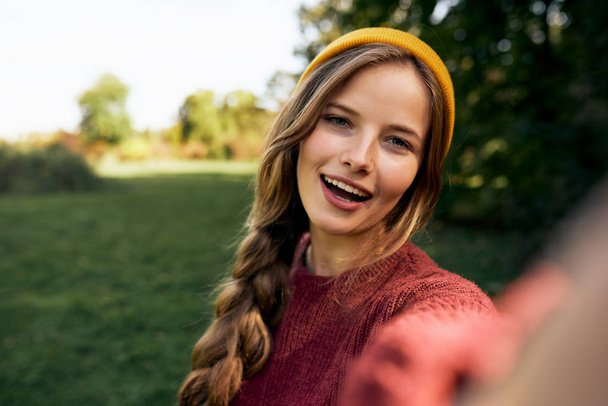 Close-up self portrait of beautiful young blonde woman smiling broadly, wearing sweater and hat, posing on nature background in the park. Happy female taking selfie. People, travel, lifestyle concept. - Zdjęcie, obraz